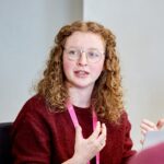 Eleanor Curry is sat at a table in front of a laptop. Her hands are raised in front of her as she addresses stakeholders who are sat around the table. The stakeholders are just out of shot of the photo.