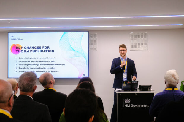 A man in a blue suit stands at the from of a seated audience, presenting a slide about the trust framework.